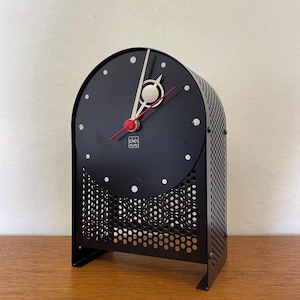Può includere: Orologio da scrivania in metallo nero con quadrante semicircolare e corpo traforato. Il quadrante è nero con punti bianchi che segnano le ore e lancette rosse e bianche. L'orologio ha il testo "deh studio".