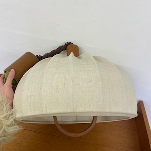 May include: A cream-colored, dome-shaped lampshade with a wooden handle. The shade is made of a textured fabric and has a wooden finial. A brown cord and a wooden fixture are visible above the shade.