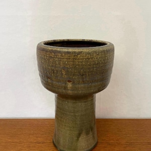 Może przedstawiać: Ceramiczny wazon na postumencie z szeroką, zaokrągloną czaszą i wąską, cylindryczną podstawą. Zewnętrzna powierzchnia pokryta jest teksturowanym, oliwkowozielonym szkliwem z subtelnymi brązowymi plamkami i poziomymi żłobieniami na czaszy.