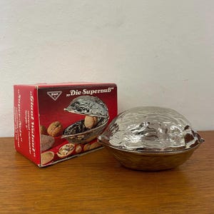 Könnte beinhalten: Ein silberfarbener Nussknacker in Walnussform mit passender Schachtel. Die Schachtel ist rot und trägt die Aufschrift "Giant Walnut" und "Die Supernuß". Der Nussknacker ist zum Knacken von Nüssen bestimmt und ein Küchenwerkzeug.