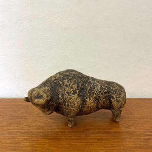 May include: A small, textured sculpture of a bison. The bison is brown with darker brown markings, and has a rough, weathered appearance. The sculpture is on a wooden surface. The sculpture is a decorative item.