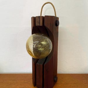 May include: A vertical dark brown wooden lamp base with a carved center section holding a spherical glass light bulb with a gold-flecked finish. A tan rope handle is attached to the top, and two small black circular knobs are visible on the side.