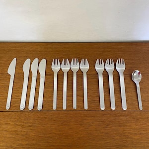 Può includere: Un set di posate in acciaio inossidabile, tra cui coltelli, forchette e un cucchiaio, disposte su una superficie di legno. Le posate hanno un design semplice e moderno con un piccolo emblema circolare sulle lame dei coltelli.