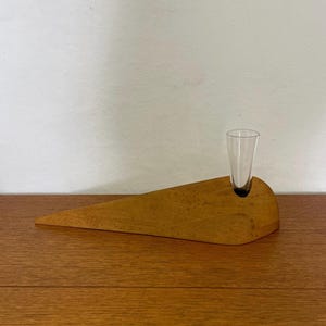 Peut inclure: Un objet en bois en forme de coin avec un petit récipient en verre transparent. Le bois est de couleur brun clair et a une finition lisse. Le verre est cylindrique et se tient droit dans un trou dans le bois.