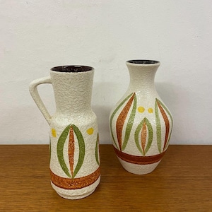 Puede incluir: Dos jarrones de cerámica con un acabado blanco texturizado y diseños geométricos en verde, naranja y amarillo. El jarrón de la izquierda tiene un asa.