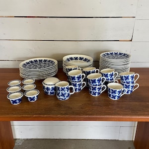Könnte beinhalten: Ein Vintage-Set aus blau-weißem Geschirr mit Blumenmuster, bestehend aus Tellern, Tassen, Untertassen, einem Milchkännchen und Eierbechern. Das Set ist auf einem Holztisch arrangiert.