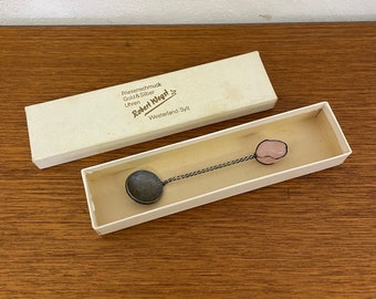 May include: A small, silver spoon with a pink glass bead on the end, in a white box. The box has the text "Robert Wegs" and "Westerland-Sylt" printed on it.