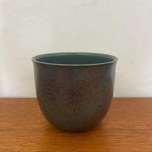 Puede incluir: Una taza de cerámica con un acabado moteado y jaspeado. El exterior es de un verde apagado con motas marrón rojizo, y el interior es de un verde más oscuro y liso. La taza está sobre una superficie de madera.