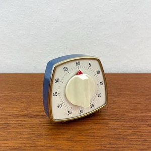 Può includere: Un timer da cucina retrò blu e bianco con quadrante color crema e indicatore rosso. Il timer ha una finitura dorata e mostra numeri da 0 a 60. Un gadget da cucina vintage.