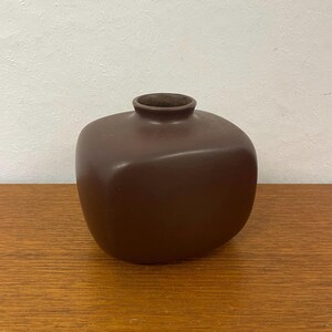 Peut inclure: Un vase en céramique marron avec une forme carrée arrondie et une petite ouverture. Le vase est de couleur marron uni et mat et repose sur une surface en bois. Le vase mesure environ 15 cm de haut.