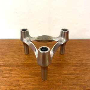 Può includere: Un portacandele in metallo color argento caratterizzato da una base a treppiede con tre gambe distinte e tre aperture circolari sulla parte superiore per le candele. La struttura è collegata da bracci metallici curvi e lisci che formano una forma triangolare.