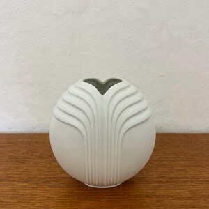 Puede incluir: Un jarrón blanco y esférico con un diseño único. El jarrón presenta unas crestas verticales y curvas que crean un patrón en forma de abanico. La abertura en la parte superior es de color gris oscuro. El jarrón está sobre una superficie de madera.