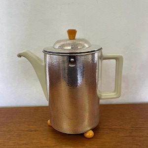 Könnte beinhalten: Eine Vintage-Kaffeekanne aus silberfarbenem Metall mit gehämmertem Muster. Sie hat einen hellbeigen Griff, Ausguss und Deckelakzente. Die Kanne steht auf drei kleinen, runden, gelben Füßen und hat einen passenden gelben Deckelknauf.