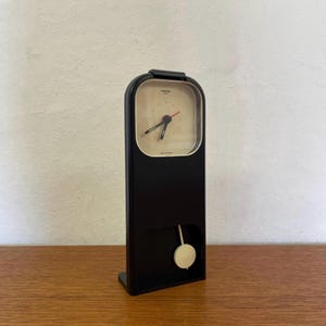 Può includere: Un orologio a pendolo nero con quadrante bianco e lancette nere. L'orologio è su una superficie di legno.