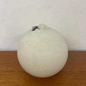 Op de afbeelding: Een ronde, gebroken witte keramische vaas met een getextureerd oppervlak. De vaas heeft een kleine, onregelmatige opening aan de bovenkant. De vaas staat op een houten oppervlak.