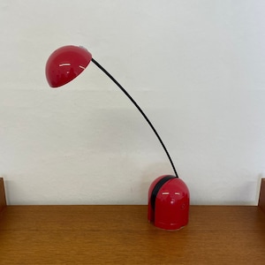 Op de afbeelding: Een rode bureaulamp met een flexibele arm en een ronde, koepelvormige kap. De lamp staat op een houten oppervlak.