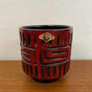 Op de afbeelding: Een rood-zwarte keramische plantenbak met een geometrisch ontwerp. De cilindrische plantenbak heeft een getextureerd oppervlak en een zwart interieur. Een kleine sticker met de tekst "Uisen Jacobs" is zichtbaar aan de voorkant.