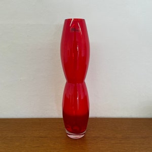 May include: A tall, red glass vase with a unique, hourglass-like shape. The vase is made of clear glass with a red tint.