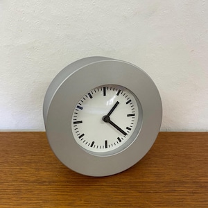 Può includere: Un orologio analogico rotondo color argento con quadrante bianco e lancette e segni neri. L'orologio è appoggiato su una superficie di legno. Il design dell'orologio è minimalista e moderno.