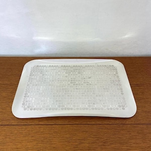 Peut inclure: Un plateau rectangulaire blanc aux coins arrondis. La surface du plateau est recouverte d'un motif détaillé et répétitif dans une nuance plus claire de blanc. Le plateau est posé sur une surface en bois.