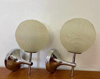 Op de afbeelding: Een paar wandlampen met bolvormige, matglazen kappen met een wervelend, gestructureerd patroon. Elke kap wordt ondersteund door een arm van gepolijst chroommetaal en een ronde, verchroomde muurbevestigingsplaat voor montage.