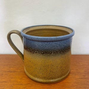 Könnte beinhalten: Eine handgefertigte Keramiktasse mit strukturierter Oberfläche. Die Tasse zeigt einen Farbverlauf, der von einem goldgelben Sockel zu einem dunkelblauen Rand übergeht. Der Griff hat einen passenden Erdtone.