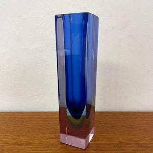 Könnte beinhalten: Eine hohe, rechteckige Glasvase mit einem tiefblauen Innenkern, der von klarem Glas umschlossen ist. Der Boden ist dick und massiv und geht in eine vertikale, säulenartige Struktur mit glatten, flachen Seiten und scharfen Kanten über.