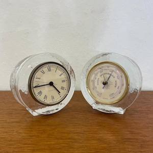 Puede incluir: Dos piezas decorativas de vidrio transparente. Una es un reloj con números romanos y la palabra "QUARTZ". La otra es un barómetro con un borde dorado y una esfera crema. Ambas tienen forma de esfera de vidrio texturizado.