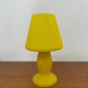 Könnte beinhalten: Eine leuchtend gelbe Tischlampe mit einem pilzförmigen Lampenschirm. Die Lampe hat einen runden Sockel und einen geschwungenen Stiel, der Schirm ist trapezförmig. Die Lampe besteht aus einem festen Material und steht auf einer Holzoberfläche.