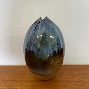 May include: A ceramic vase with a unique shape and a glossy finish. The vase has a dark blue and brown color scheme with a textured surface. The top of the vase has several slits, adding to its distinctive design.