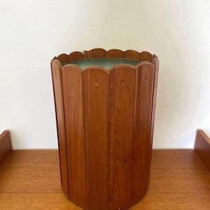 May include: A cylindrical wastebasket constructed from vertical wooden slats with a warm brown finish. The top edge features a scalloped design, and small metal fasteners are visible along the length of each slat, securing the structure.