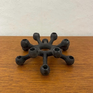 Può includere: Un portacandele nero a forma di stella con otto vani circolari per candele. Il portacandele è realizzato in un materiale nero opaco e poggia su una superficie di legno. Il design è moderno e minimalista.