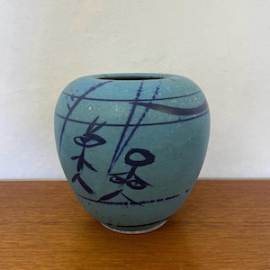 Könnte beinhalten: Eine blaue Keramikvase mit einem stilisierten Schwarz-Weiß-Design aus zwei Figuren mit Augen. Die Vase hat eine abgerundete Form und eine glatte, matte Oberfläche.