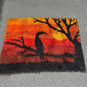 May include: Rectangular shag rug with a sunset scene. The rug features a gradient of orange, red, and black, with a black silhouette of a bird perched on a branch and a tree. The rug measures approximately 36 inches by 24 inches.