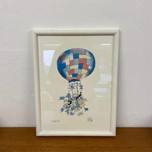 Puede incluir: Una impresión de dibujos animados enmarcada de un globo aerostático con un grupo de personas colgadas del cesto. El globo es multicolor con cuadrados azules, rojos, rosas y blancos. Las personas están dibujadas en un estilo caprichoso con cuerpos azules y caras blancas. La impresión está firmada "Stein" en la esquina inferior derecha y "05/100" en la esquina inferior izquierda.