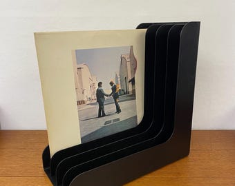 Puede incluir: Soporte negro para discos de vinilo con un diseño curvo. El soporte muestra la portada de un álbum con dos hombres dándose la mano frente a un edificio. El soporte es de material oscuro y se apoya sobre una superficie de madera.