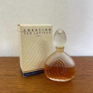 Könnte beinhalten: Eine durchsichtige Glasflasche von Creation Ted Lapidus Paris Eau de Toilette. Die Flasche hat ein strukturiertes, wellenförmiges Design und einen mattierten Glasverschluss. Die Flasche steht auf einer Holzoberfläche.