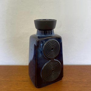Könnte beinhalten: Eine dunkelblaue Keramikvase mit schwarzem Deckel und zwei Spiralmustern. Die Vase hat eine rechteckige Form mit abgerundeten Kanten und steht auf einer Holzoberfläche.