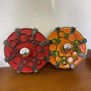 May include: Two decorative ceramic wall sconces. One is red with a silver orb and dark accents, the other is orange with green and dark accents. Both have a circular design with a central orb.