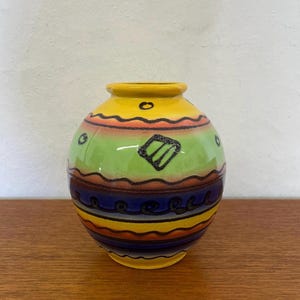 Puede incluir: Un jarrón de cerámica amarillo, verde, azul y naranja con un diseño geométrico. El jarrón tiene una forma redondeada y una abertura estrecha.