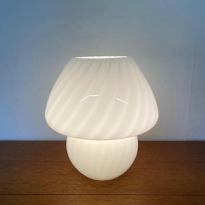 Könnte beinhalten: Eine weiße Tischlampe in Pilzform mit einem Wirbeldesign auf dem Schirm. Die Lampe hat einen abgerundeten Sockel und einen pilzkappenförmigen Aufsatz. Die Lampe ist aus Glas und steht auf einer Holzoberfläche.