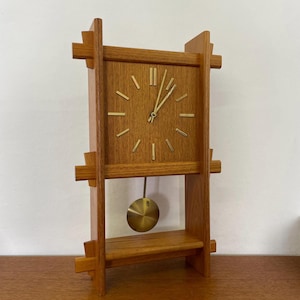 May include: A wooden mid-century modern clock with a square face and brass hands and pendulum. The clock has a light brown finish and a shelf below the clock face.