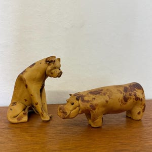 May include: Two ceramic animal figurines. One is a seated dog with a tan and brown mottled pattern. The other is a hippo with a similar colour pattern. Both are on a wooden surface.