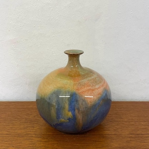 Peut inclure: Un vase rond en céramique avec un col étroit. Le vase présente un mélange de couleurs, notamment orange, bleu et vert, créant un effet marbré. Le vase est posé sur une surface en bois.