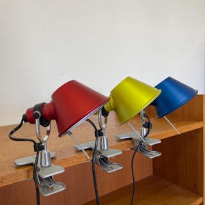 Könnte beinhalten: Drei farbige Klemmleuchten mit roten, gelben und blauen Schirmen. Jede Lampe hat einen silberfarbenen Klemmfuß und ein schwarzes Kabel. Die Lampen stehen auf einem Holzregal und bieten einen lebendigen Akzent.