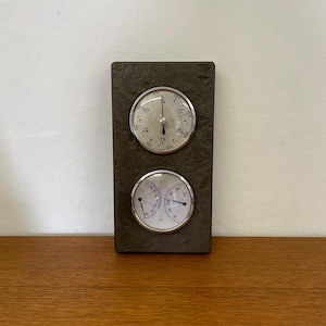 May include: A gray slate rectangular wall mounted thermometer and barometer with two round analog gauges. The top gauge is a barometer with a needle pointing to 76. The bottom gauge is a thermometer with a needle pointing to 20 degrees Celsius.