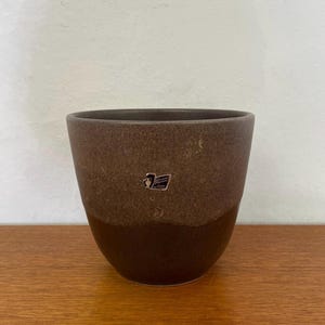 Puede incluir: Una maceta de cerámica marrón con un diseño de dos tonos. La parte superior tiene una textura moteada, mientras que la sección inferior es de color marrón oscuro sólido. Un pequeño logotipo es visible en la parte delantera. La maceta está sobre una superficie de madera.