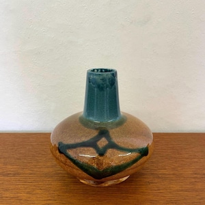 Może przedstawiać: Ceramiczny wazon z szerokim, zaokrąglonym korpusem i wąską szyjką. Podstawa wazonu pokryta jest nakrapianym brązowo-beżowym szkliwem, podczas gdy szyjka ma głęboki, morski odcień niebieskiego. Ciemnozielony, geometryczny wzór zdobi korpus naczynia.