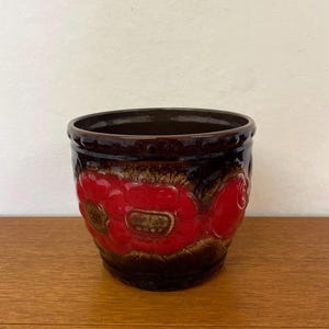 Peut inclure: Un pot de fleurs en céramique avec un extérieur marron foncé et un intérieur brillant. Le pot présente des fleurs rouges en relief avec des accents dorés. Le pot est posé sur une surface en bois.