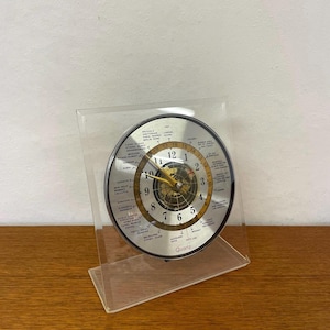 Può includere: Un orologio da mondo al quarzo argentato e dorato con un supporto in acrilico trasparente. Il quadrante dell'orologio presenta una mappa del mondo e mostra l'ora con lancette dorate. L'orologio è contrassegnato "Quartz".